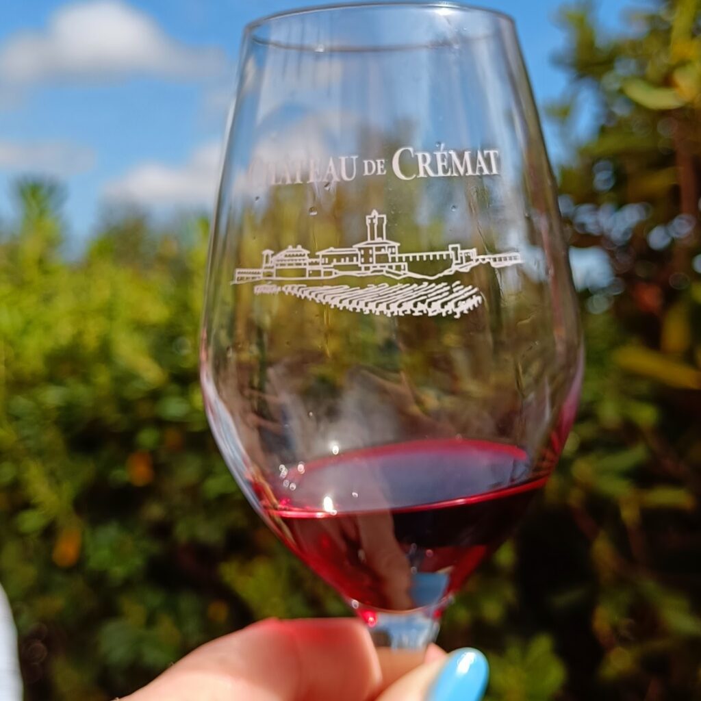 A glass of wine held up to the sky, holding a small measure of red wine. The glass reads "Chateau de Cremat"